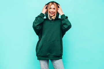 Young beautiful smiling female in trendy summer green hoodie. Sexy carefree woman posing near blue...