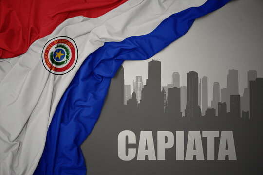 abstract silhouette of the city with text Capiata near waving national flag of paraguay on a gray background.