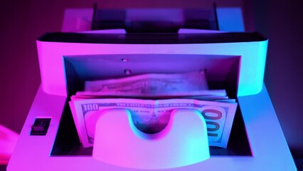 A woman's hand puts 100 USA dolars bills into the banknote detector counter. The money counting machine processes the money bundle. Equipment for counting money. Financial crimes. - Powered by Adobe