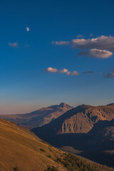 Naklejka premium Vertical view of bare mountain peaks about tree line as seen from trail ridge road in Rocky Mountain National Park, Colorado, at sunset; blue sky with beautiful clouds and rising moon in background