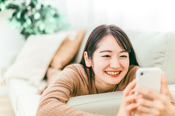 家のソファでスマホを使う日本人女性（リラックス・笑顔）
