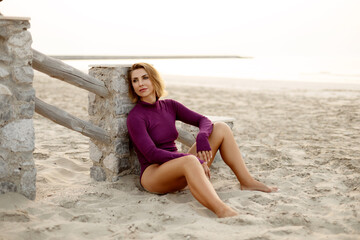 A glamorous young woman in a purple bodysuit is sitting on the sand on the beach