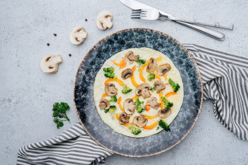 Omlette with mushrooms, paprika, broccoli on a plate. On a napkin on a table. Decorated with champignons halves and cutlery.