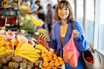 Portrait of a young smiling woman buying fruits at the local market. Shopping with reusable mesh bag. Sustainability and organic food concept