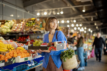 Portrait of young cheerful woman holds box full of fresh fruits and berries at local market. Woman buying food at market. Concept of traditional offline retail of local products