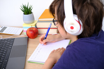 smart child in headphones, boy 9-10 years, primary school student writes in front of open laptop, doing homework, Back to school concept, doing homework, practicing spelling skills