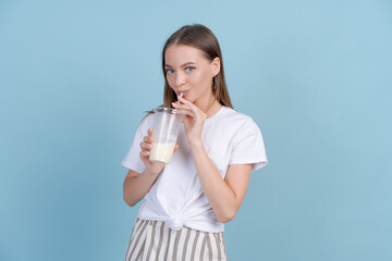 Cute caucasian young girl in white t-shirt and striped pants drinks milkshake on blue background and smiles with her teeth while looking at the camera