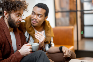 Gay couple with different nationality having close conversation while sitting on a couch at home....