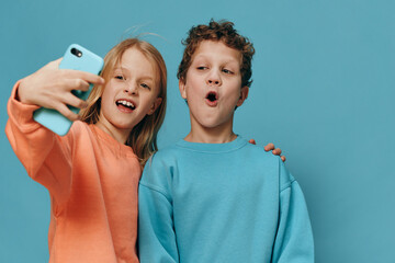 cute, beautiful children, brother and sister stand in bright clothes on a blue background and the girl takes a selfie on her smartphone hugging her brother