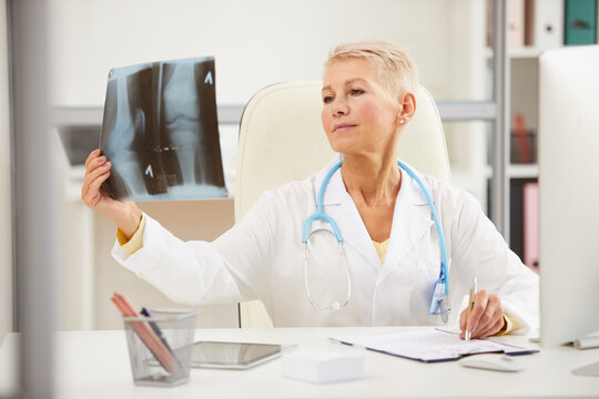 Pensive attractive mature radiologist with stethoscope on neck sitting at table and analyzing x-ray image on knee
