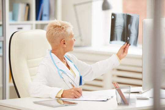 Qualified mature female orthopedist in lab coat sitting at table and examining x-ray image of knee