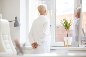 Pensive female director of Medicine wearing white coat holding hands behind back and looking out window while thinking of solution