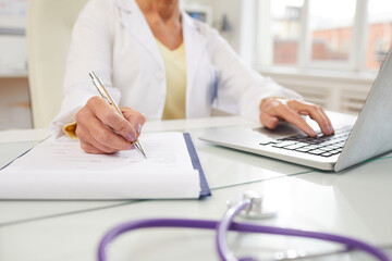Close-up of unrecognizable doctor sitting at table and using laptop while filling document with medical history of patient