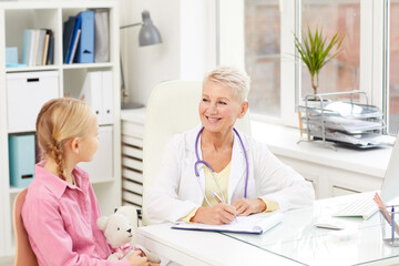 Obraz premium Positive friendly female pediatrician with short hair sitting at table and filling medical history while talking to child in office