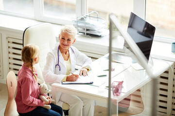 Obraz premium Smiling friendly mature blond-haired doctor with stethoscope on neck sitting at desk in office and talking to little patient with toy