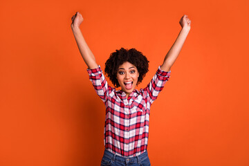Portrait of astonished overjoyed girl raise hands fist up success isolated on orange color background