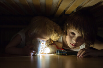 Little child, hiding under the bed, hugging teddy bear and holding flashlight,