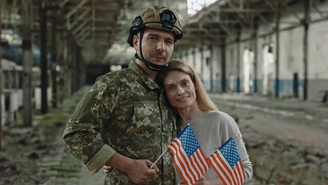 Portrait of male warrior in military uniform hugging lovely wife while standing at destroyed factory. Young family looking at camera with smile and holding american flag in hands.