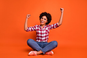 Full size portrait of astonished positive girl sit floor raise fists celebrate achieve isolated on orange color background