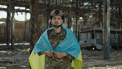 Front view of fearless soldier in military uniform and helmet covered his shoulder with Ukrainian flag. Patriotic male hero defending motherland during war. Background of destroyed steel plant.