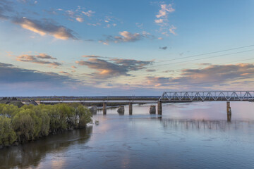 Evening landscape, sunset on the river. Spring, green trees in the foreground. View of the industrial area of the railway bridge. Outskirts.