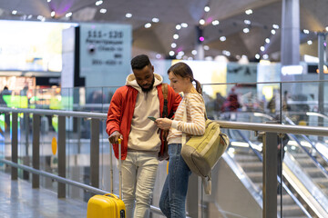 Young european woman with smartphone showing direction to african man traveler in airport. Couple of friends travelers looking at mobile phone screen while traveling together, searching location