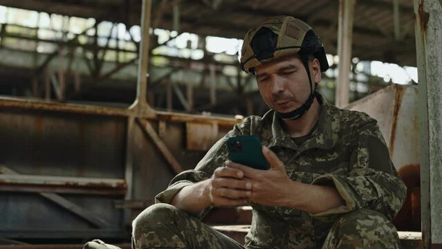 Smiling man in military uniform using modern smartphone while sitting near burnt bus on destroyed factory. Army officer looking through family photos on mobile phone.