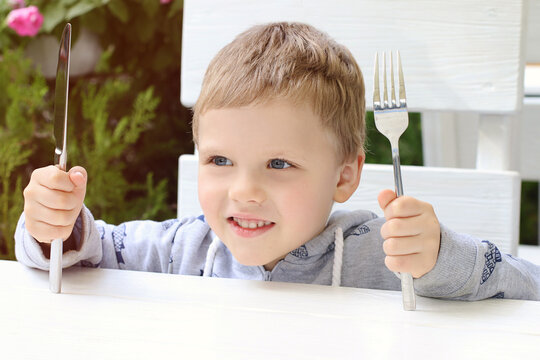 The smiling child shows that he wants to eat. Kid holding fork and knife over the table. The interior in a modern restaurant outdoor.