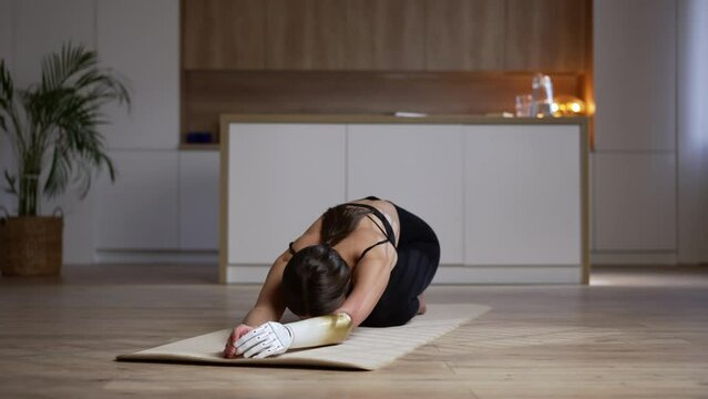 Young woman with bionic arm is doing yoga and staying at home during lockdown spbas. 4k Attractive lady leans on mat and does exercise or asana, poses for camera and trains slowly in room. Caucasian