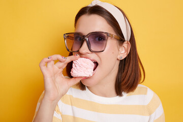 Image of joyful Caucasian young adult woman biting dessert looking at camera with satisfied expression, biting marshmallow, wearing casual clothing, posing isolated over yellow background.