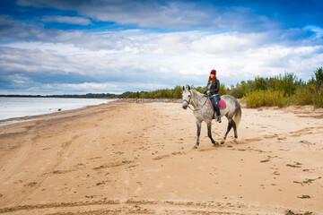 wearing jeans, jacket and warm hat female horseback raider goes on horseback grey horse along seashore