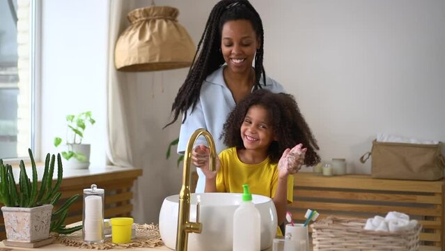 Young mother, cute daughter are doing daily routine and having fun in home bathroom spbd. Close view of american african woman, girl wash hands thoroughly and use soap, talk with smiles and stand near