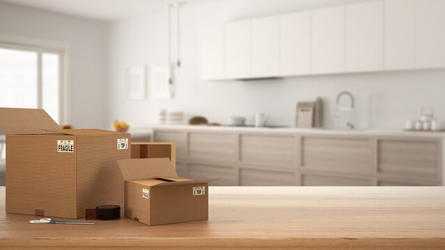 Wooden table, desk or shelf with stack of cardboard boxes over blurred view of scandinavian kitchen with appliances, modern interior design, moving house concept with copy space