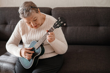 Elderly woman 70+ sits on the couch at home and plays ukulele. Adaptation of pensioners in the modern world. New knowledge and skills, hobby. Prevention of Alzheimer's disease, sclerosis or dementia