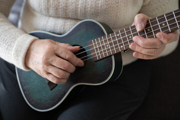 Hands of an elderly woman 70+ sitting on the sofa at home and playing the ukulele. Adaptation of pensioners in the modern world. New knowledge and skills. Prevention of Alzheimer's disease, sclerosis 