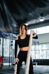 Powerful woman training battle ropes at cardio workout in dark gym. Professional athlete exercise fitness sport club equipment. Strong bodybuilder lifting weights. Athletic person effort