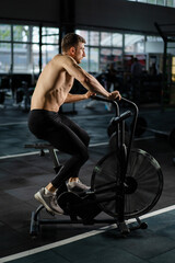 Fototapeta na wymiar Young Athletic middle aged man training on air resistance bike, cross training workout set in gym. Active man spinning a air bike in gym with trainers. Male training on air bike.