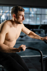 Obraz premium Young Athletic middle aged man training on air resistance bike, cross training workout set in gym. Active man spinning a air bike in gym with trainers. Male training on air bike.