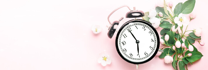 Black alarm clock that shows 7 o'clock on a pink background with apple tree spring summer flowers...