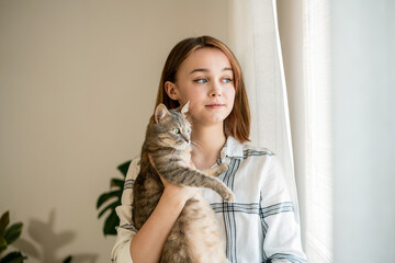 Indoor shot of amazing lady holding cat . Portrait of young woman holding cute striped cat with green eyes and looking at the window. Female hugging her cute kitty. Adorable domestic pet concept.
