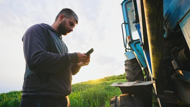 Man in field with phone in his hands stands by his tractor and knocks on wheel, checking condition of machine to diagnose serviceability of equipment. Running a small business selling grown products.