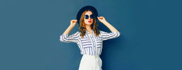 Portrait of beautiful young woman wearing white striped shirt, black round hat on dark blue background