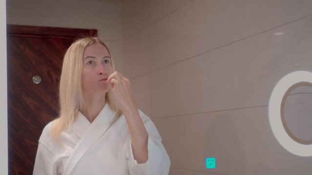 Beautiful young woman in white coat or pajamas brushes her teeth in front of mirror in bathroom. Mirror image of beautiful woman washing her teeth is daily concept of oral hygiene. Fresh good morning