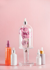 Cosmetic bottles with orchid flowers in bell jar at pale pink background. Facial treatment concept...