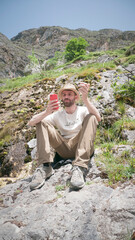 Hombre blanco sentado en ladera de montaña rocosa mirando su teléfono móvil