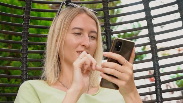 Positive European Caucasian blonde woman looks into her smartphone and swings in hanging rattan chair. Portrel girls. Business woman works in app on her phone. Touching display with your finger.