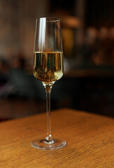 Glass of champagne standing on wooden table in soft focus on naturally blurred dark background. White wine with bubbles. Celebration, happy holidays concept