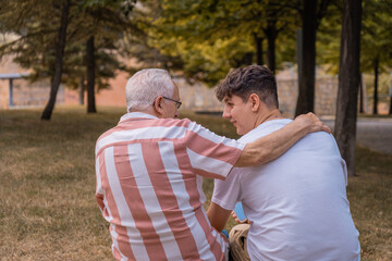 Abuelo abrazando a su nieto sentados en el parque, con expresión cariñosa. Fotografía horizontal.