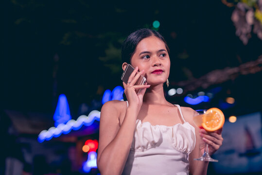 A modern and trendy young woman trying to call her late friend while drinking a glass of tequila sunrise. Vibrant nightlife outdoor scene at a bar or cafe with neon lights.