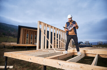 Male carpenter building wooden frame house on pile foundation. Man builder standing on construction...